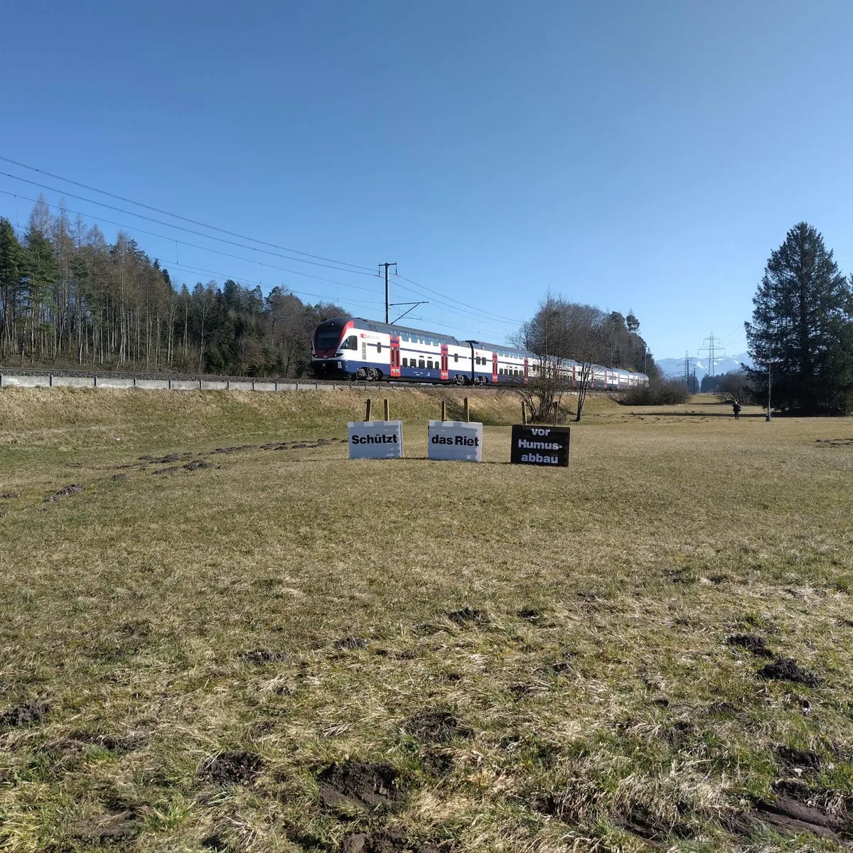 Mit diesem Schild wollten Interessensvertreter auf den Humusabbau aufmerksam machen. Man sieht ein Landstück, auf dem ein Zug fährt. Darauf stehen Schilder mit der Aufschrift: Schützt das Riet vor Hummusabbau.